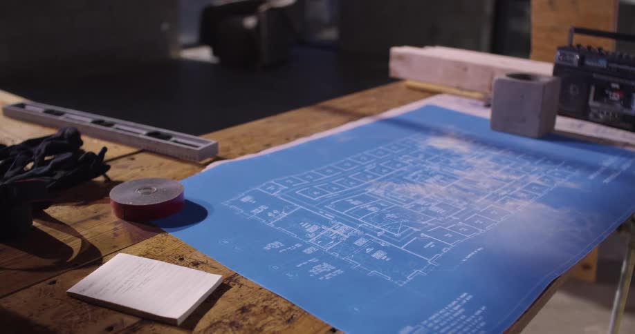 View zooming in on a set of blueprints for a building project that is laying on a wooden table surrounded by tools and construction materials.