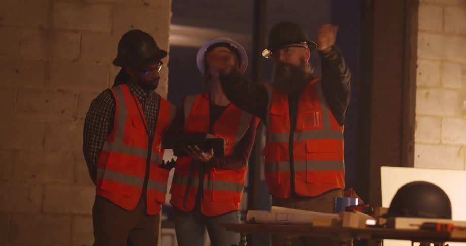 View from a distance of three construction workers, a woman and two men, discussing the plans over a tablet and gesturing to different areas in a project site full of tools.
