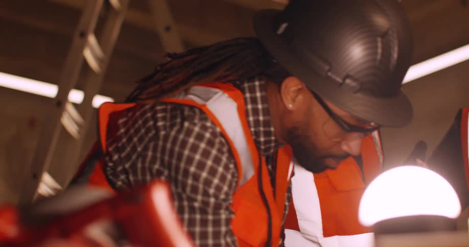 Civil engineers wearing reflective vests and hard hats examine tools and equipment, and discuss project details at an indoor construction site.