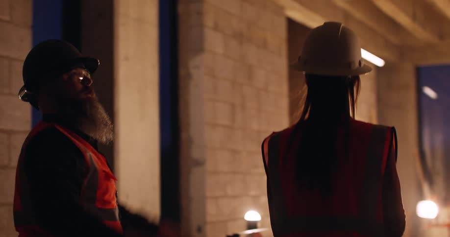 Three construction workers walk an indoor job site at night with tablet in hand and discuss the building plans as they gesture to different areas.