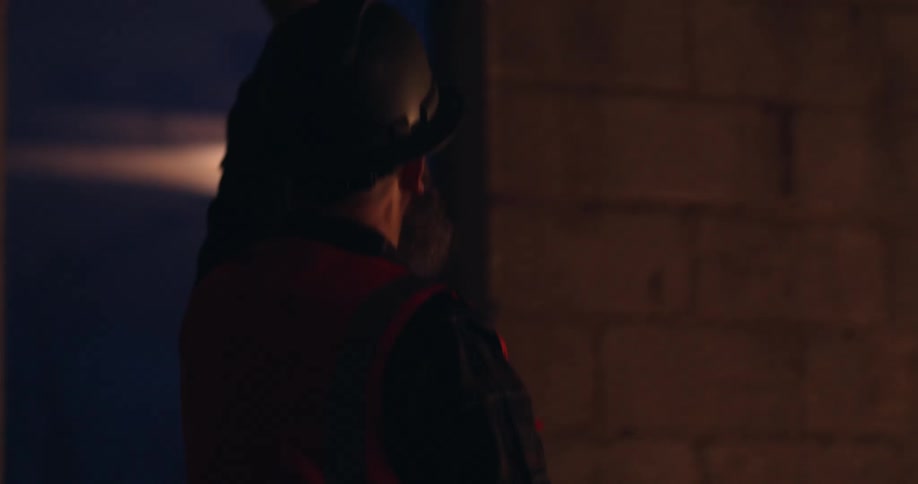 A bearded, white, male construction worker wearing a reflective vest and hard hat walks around a building site at night and gestures left and right to point out details to someone off camera.