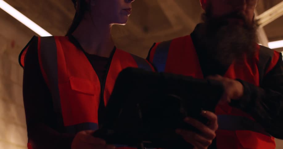 Three diverse engineers wearing hard hats and safety gear analyze information on a tablet inside a construction site at night.