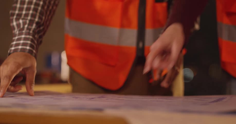 The hands of three different people wearing orange safety vests on a construction site are interacting with a blueprint on a table full of scattered tools.