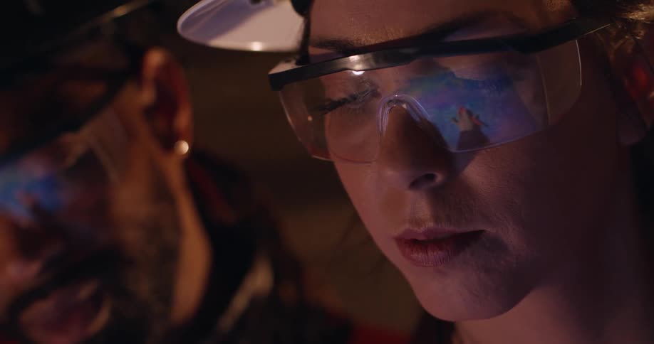 Two construction workers, a black male and a white female, wearing hard hats and safety gear look down at a screen that is reflected in their protective goggles.
