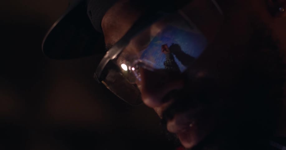Close up of a man of color wearing safety glasses and hard hat in a dark room and looking down with the reflection in his glasses of a lamp and his hand drawing on blueprints on the table below.