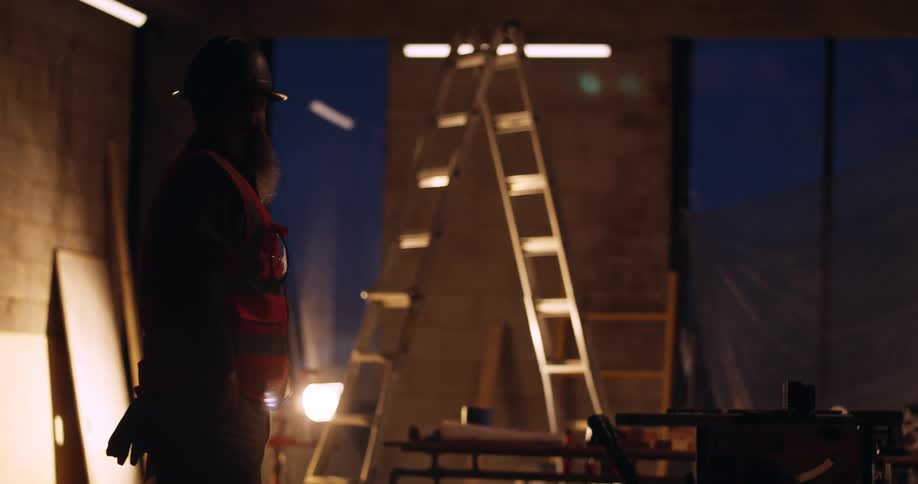 A shadowed construction worker is standing in a building under construction at night with a ladder and other tools and gear nearby lit by a floodlight.