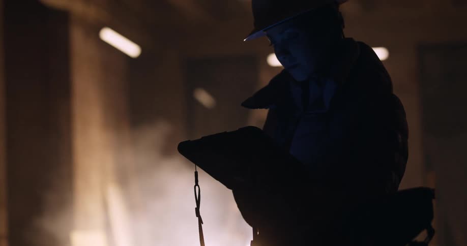 Silhouette of a female engineer in a hard hat and safety gear holding and looking down at a tablet while standing in a dusty construction site at night.