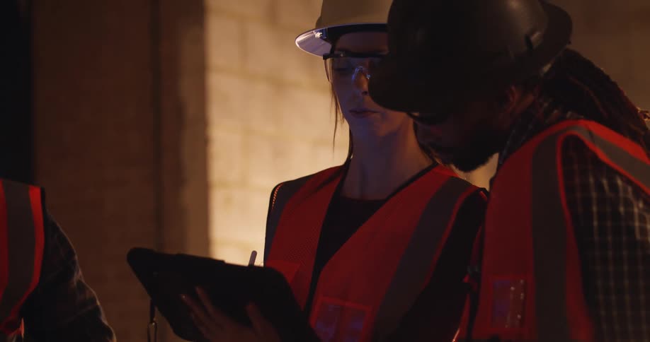 A female and two male civil engineers wearing safety gear are talking and interacting over a tablet while on a construction site at night.