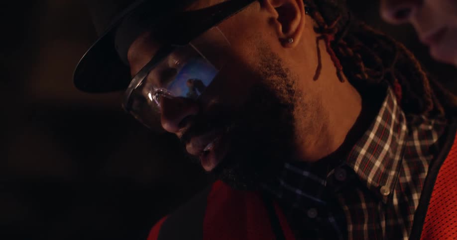 A bearded black male construction worker wearing hard hat and safety gear stands in a dark room looking down at something and speaks to a woman next to him.