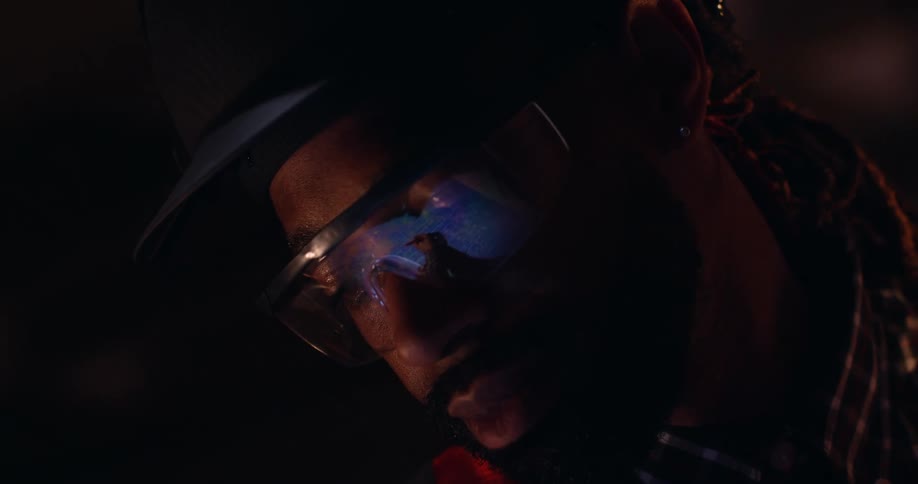 Close up view of the face of a black man wearing safety glasses and a hard hat in a dark room with the reflection in his glasses of his hand marking up blueprints on the surface below.