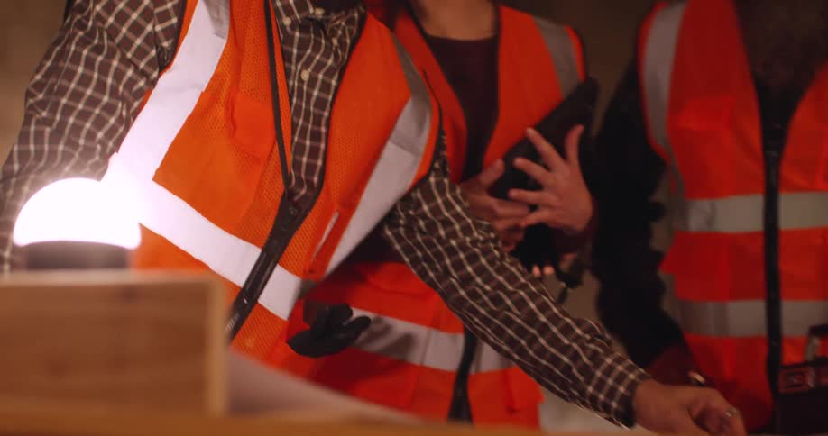 A black male civil engineer at a building site pulls a rolled up set of blueprints over to a surface and unrolls it to show two other construction workers, a man and a woman, the project plans.