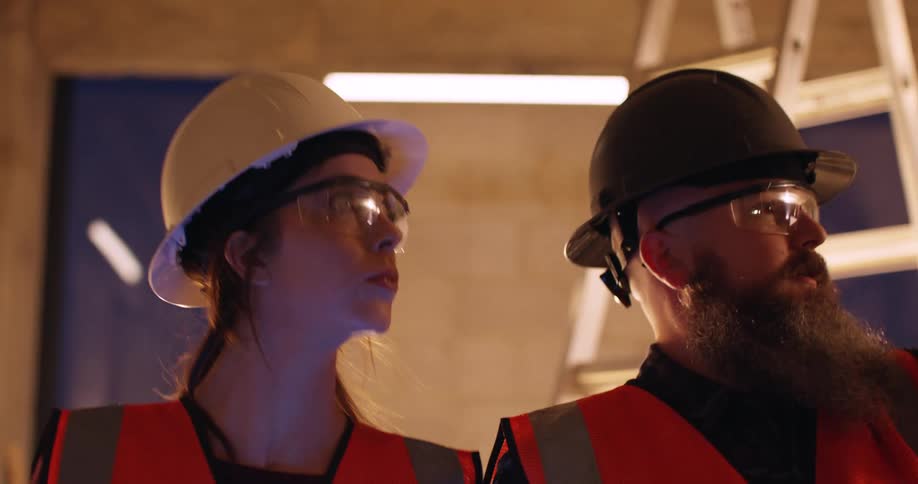 Three construction workers stand in a building site at night and they discuss project details as the two men gesture around and the woman makes notes on a digital device.