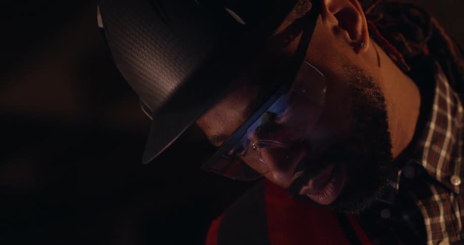 Close up of a black man in a hard hat, safety glasses and vest looking down intently at something on the surface below him.