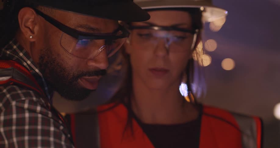 A black male engineer and a white woman, both wearing safety gear and hard hats in an active construction area, look down at something on a digital device reflected in their safety glasses as a welder creates sparks behind them.