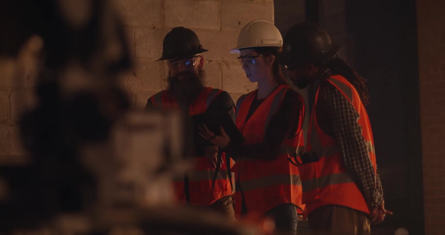 Side view from behind objects of engineers in safety gear discussing progress over a tablet in an indoor construction site at night