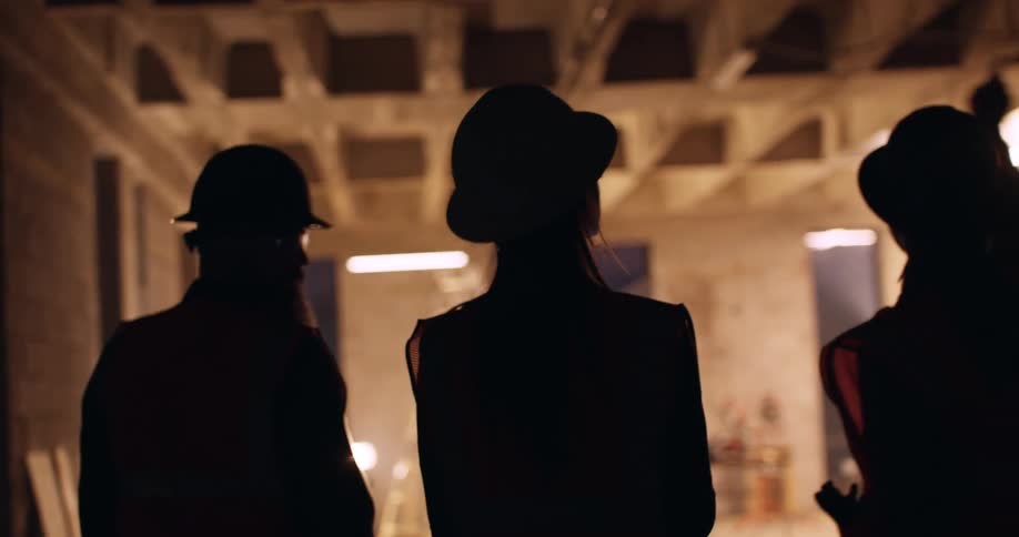 View from behind of three construction workers, wearing orange safety vests and hard hats, walking around an active construction site at night and pointing out different areas as they have a conversation.