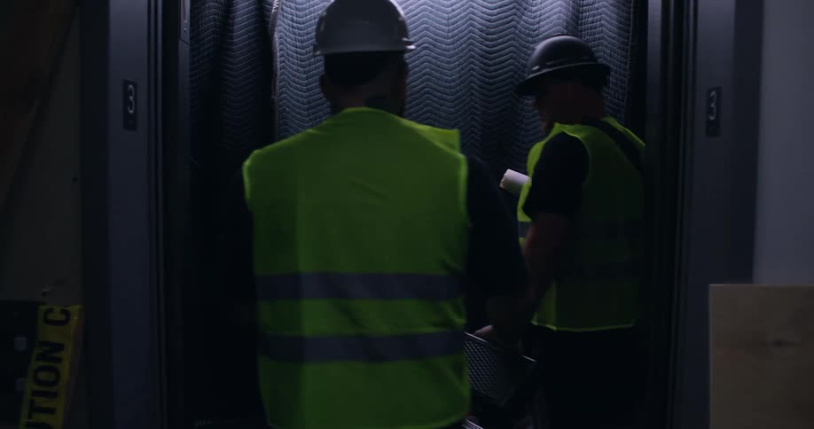 Elevator doors open to a room under construction and 3 workers in hard hats and orange safety vests get off, and then two other workers in yellow safety vests step in and press the button to another floor as the doors close.