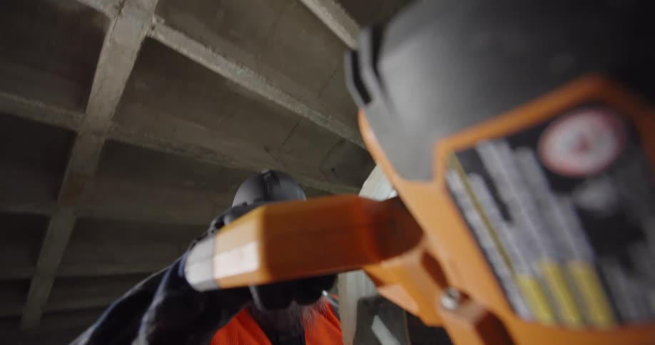 View from the handle of a miter saw at a construction site as a bearded worker pulls it down and the circulating blade makes a cut.