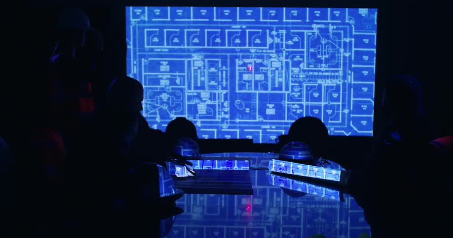 The silhouettes of a few people in a presentation room with the lights off viewing a set of blueprints being projected on a screen and one person circling details with a laser pointer.