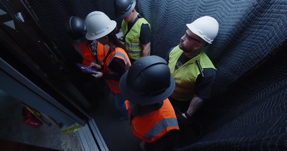 Five construction workers in hard hats and safety gear exiting a padded elevator into a building site.