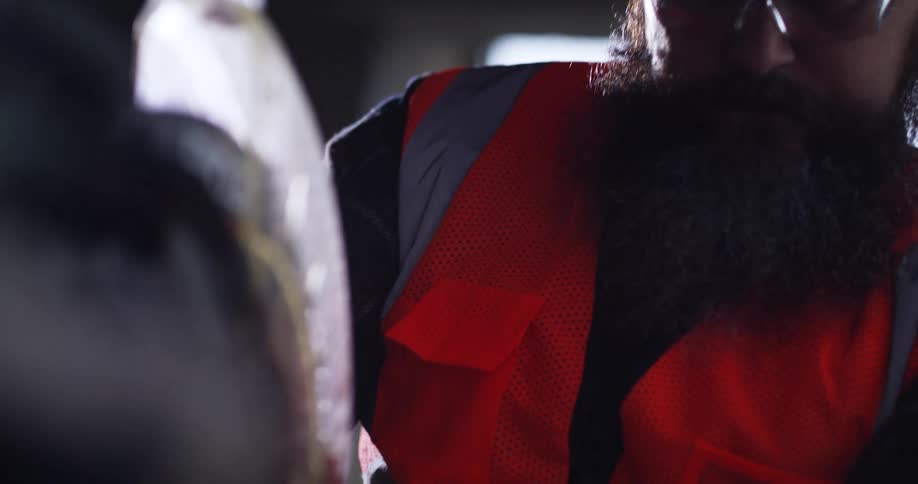Close up POV from behind a miter saw of a masculine, bearded construction worker in a hard hat and safety gear pulling the circulating blade handle down to make a cut.