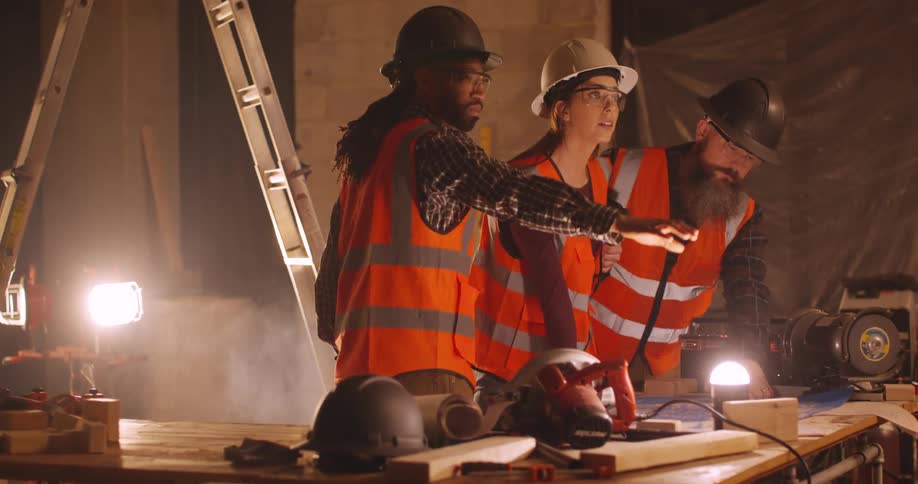 View panning from behind a miter saw of three engineers, a woman and two men, wearing hard hats and safety gear, discussing project plans on a construction site at night, illuminated by a floodlight.