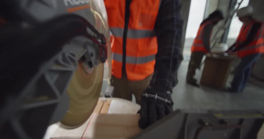 POV from the handle of a circulating miter saw of a bearded worker making a cut with the blade on a piece of wood as two other workers have a discussion behind him in an active construction site.