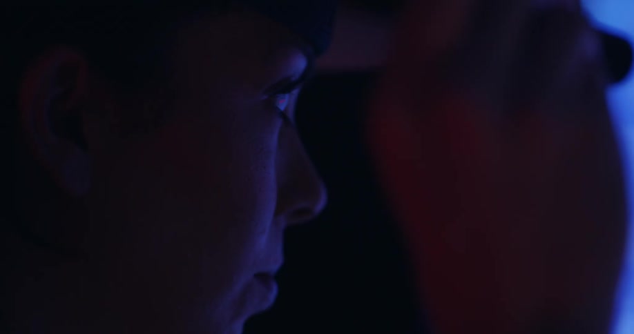 A woman in shadowed profile and wearing a hard hat is in a dark room lit only by a blue light and is holding and moving something in her hand at eye level.