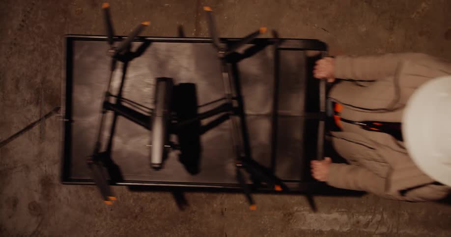 Static view from above of a person walking by wearing a white hard hat while pushing a cart with a drone on it.