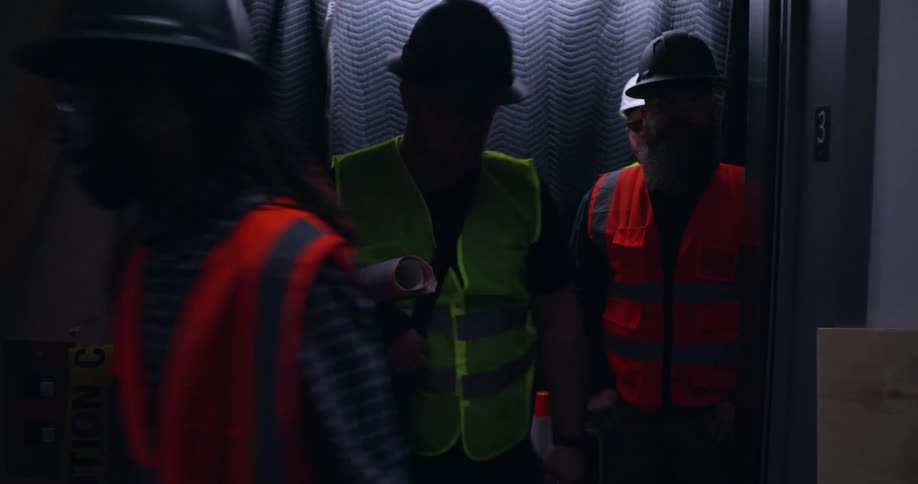 Five diverse workers wearing hard hats, protective vests, and safety glasses exit an elevator at a construction site