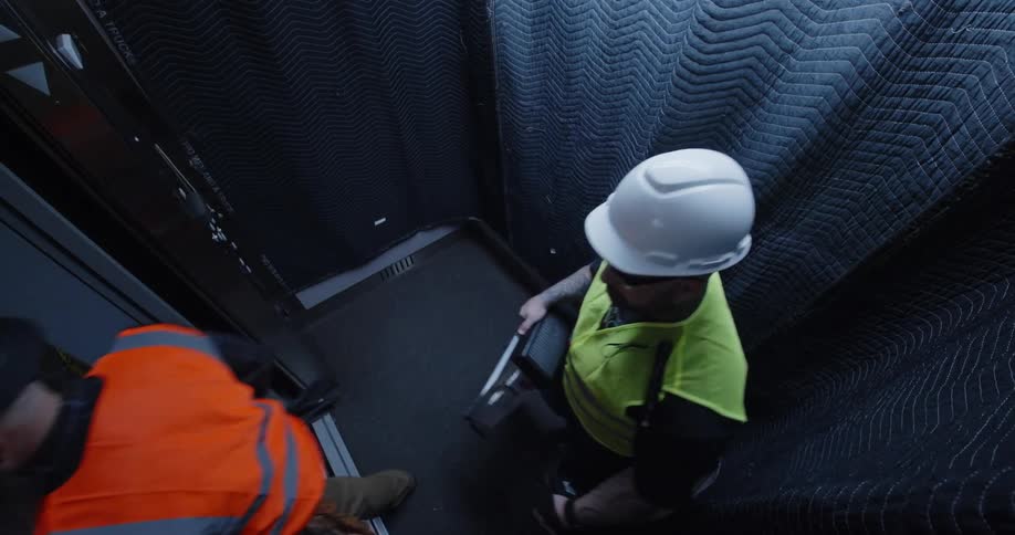 A group of civil engineers and construction workers wearing hard hats and safety vests exit a padded elevator into a room under construction.