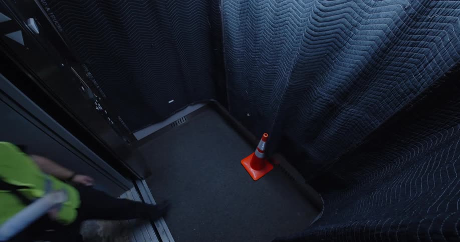 Three construction workers in an elevator wearing hard hats and orange safety vests exit when the door opens and are replaced by two other workers in yellow vests who push the button for another floor as the doors close.