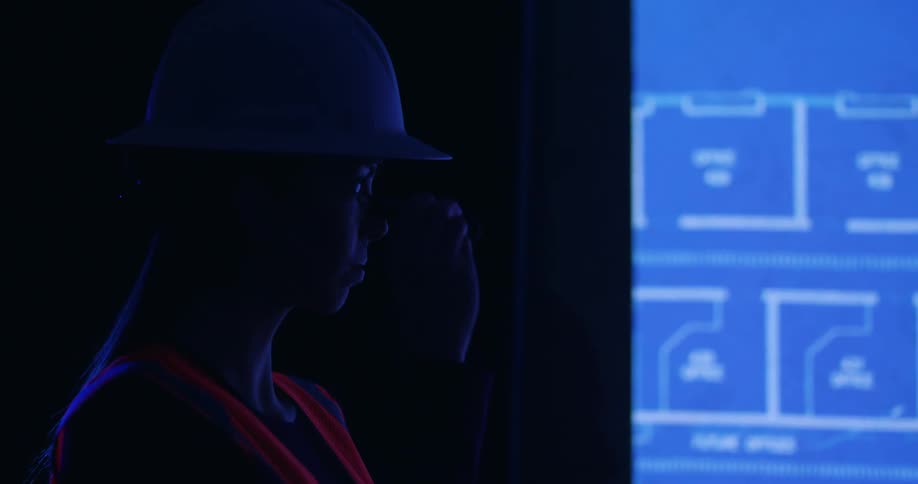 The shadowed side profile of a female civil engineer in a hard hat and safety vest standing in a dark room next to a screen projecting digitally rendered blueprints.