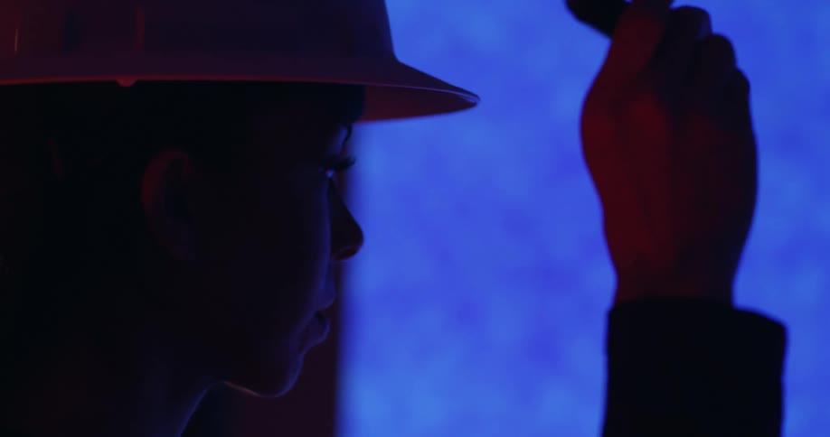 Close up of a woman in a hard hat in a dark room holding up a laser pointer toward a blueprint projector.