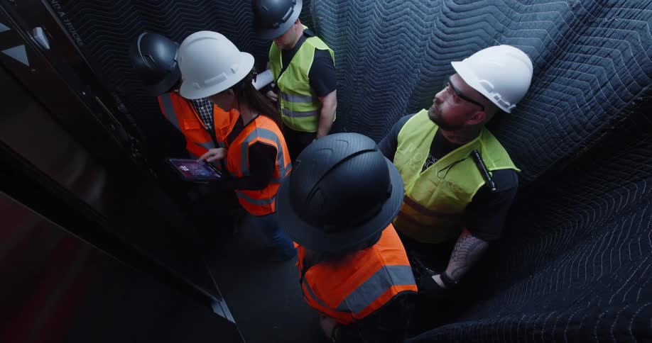Construction workers ride in a padded elevator while one of them types on an electronic tablet and talks with the person next to her, and then they all exit when the doors open.