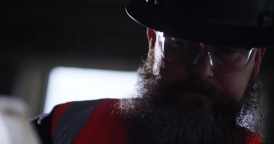 Close up of a bearded construction worker in hard hat, safety vest, and eye protection operating a miter saw.
