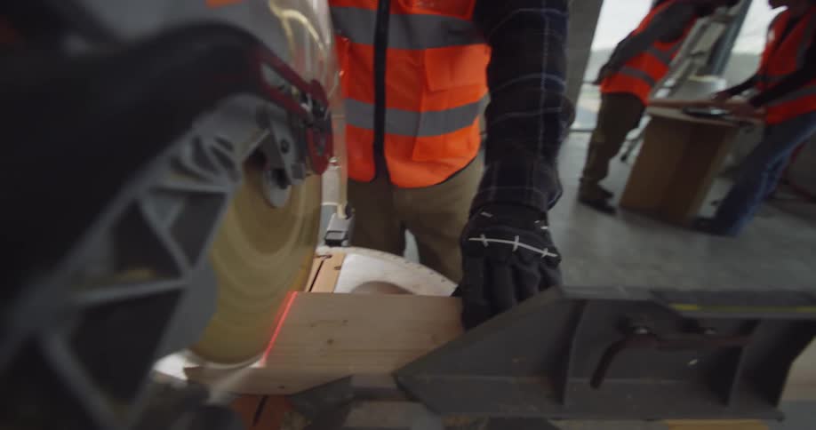 POV from the arm of a miter saw as the circulating blade is pulled down to cut a piece of wood by a bearded male construction worker at an active building site with other workers in the background.