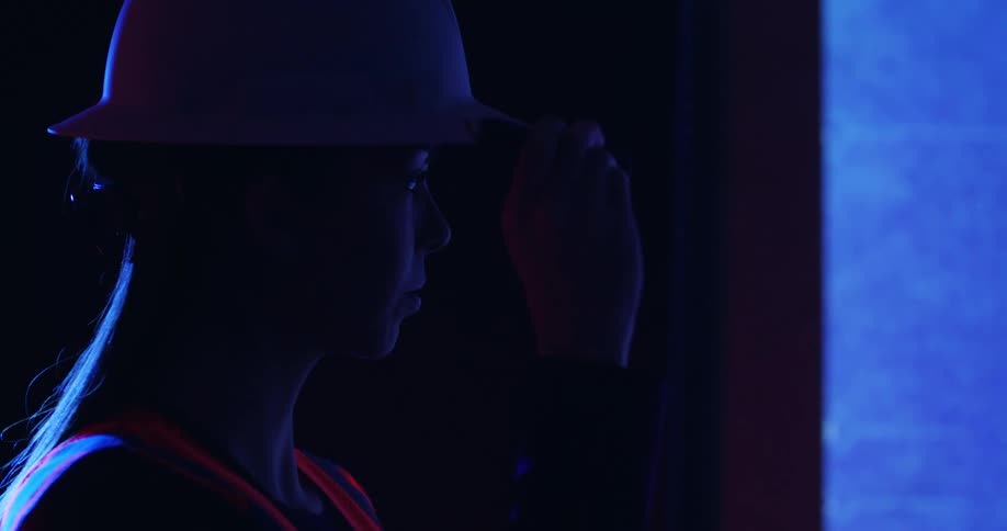 A female construction worker in shadowed profile wearing orange safety vest and hard hat stands in a room illuminated by blue light and holds up a laser pointer in her hand at eye level.