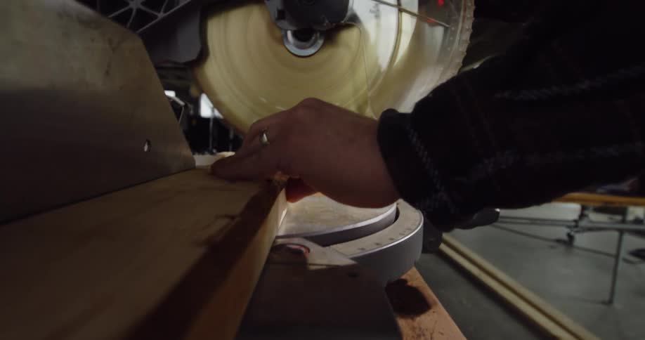 View from a piece of wood zooming in on a worker's hands using a miter saw to cut a piece of wood in a building under construction.
