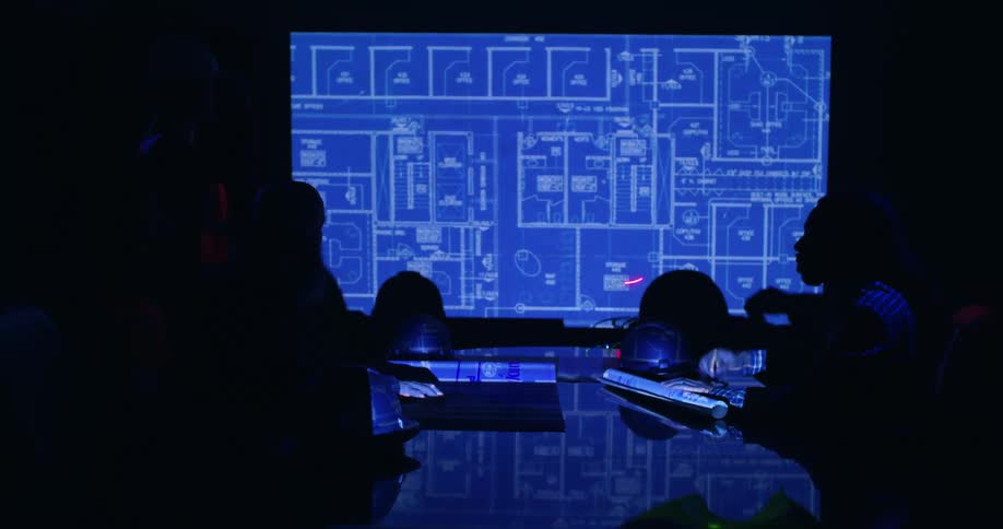 The silhouettes of a small group of people sitting at a boardroom table in a dark room viewing a blueprint on a screen and one of them circling details with a laser pointer.