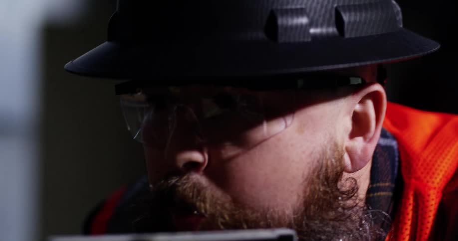 A bearded male construction worker wearing a hard hat, safety vest, and eye protection bends over to blow sawdust off of the wood board he just cut with a miter saw.