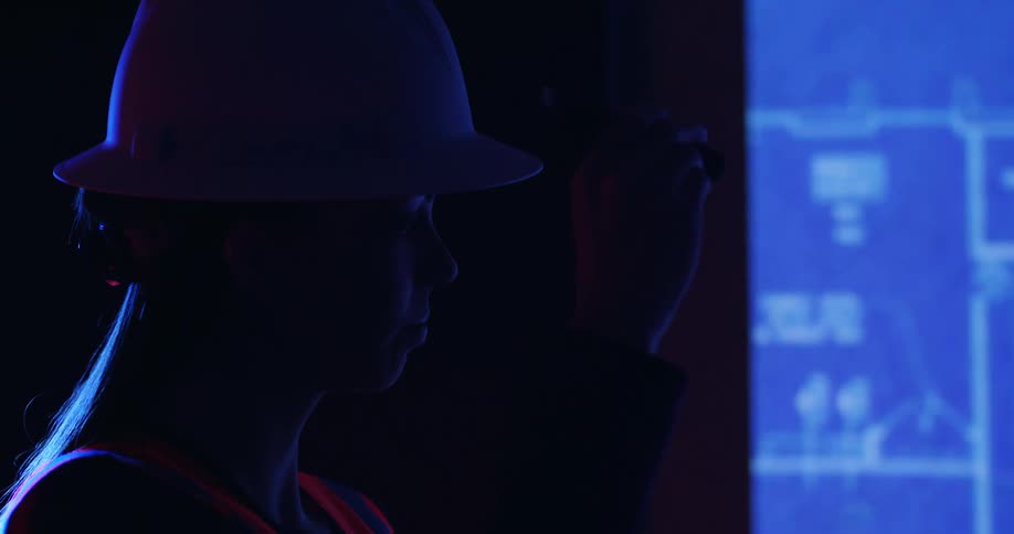 Side view of a woman in a hard hat and safety gear standing in the dark in front of a digital blueprint projector and moving the view around.