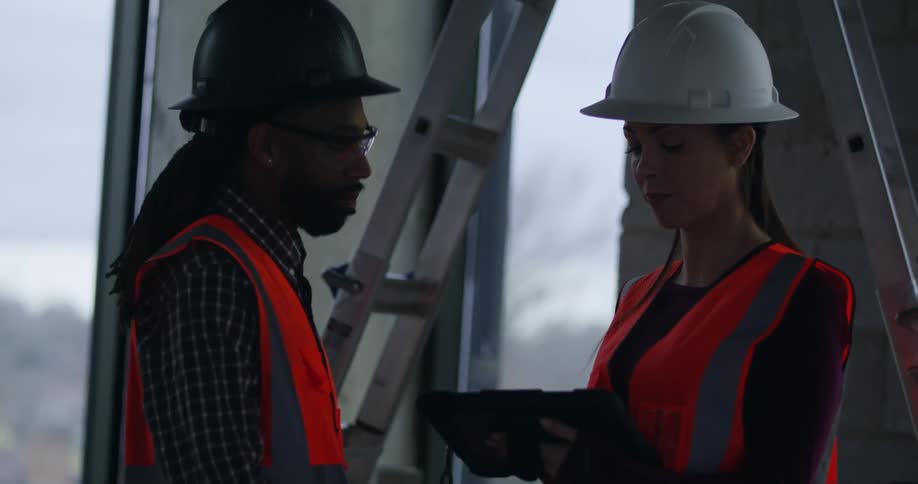 A white woman and black man in hard hats and safety gear stand talking over a tablet with a ladder behind them in a building under construction.