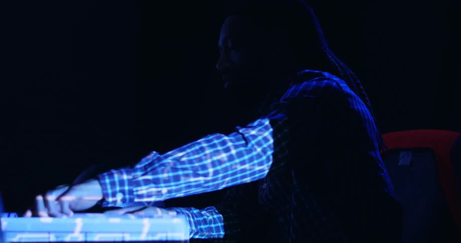 A black man sitting at a table in a dark room lit only by a projector rolls up a set of blueprints and sets them aside while speaking to someone else in the room and adjusting a hard hat on the surface.