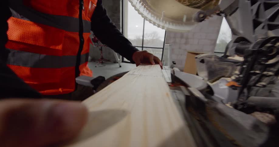 POV from a piece of wood of a bearded man with hard hat and safety gear who is working on a construction project and starts by measuring the wood then walks it over to a miter saw to cut it.