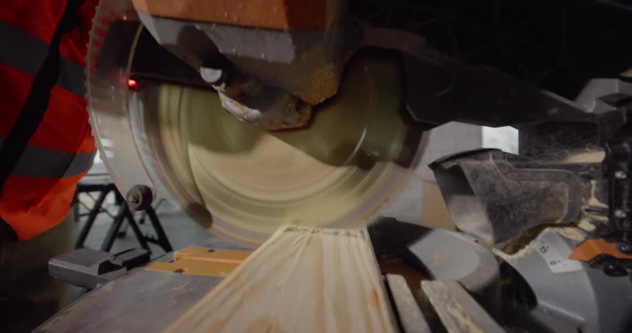 View from a piece of wood of a pair of hands operating a miter saw to cut the wood and then walk away with it.