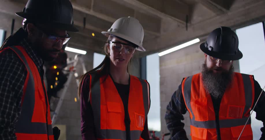 Three workers, a woman and two men, standing in a building under construction with a welder throwing up sparks on a ladder behind them as they discuss project plans they are looking down at.