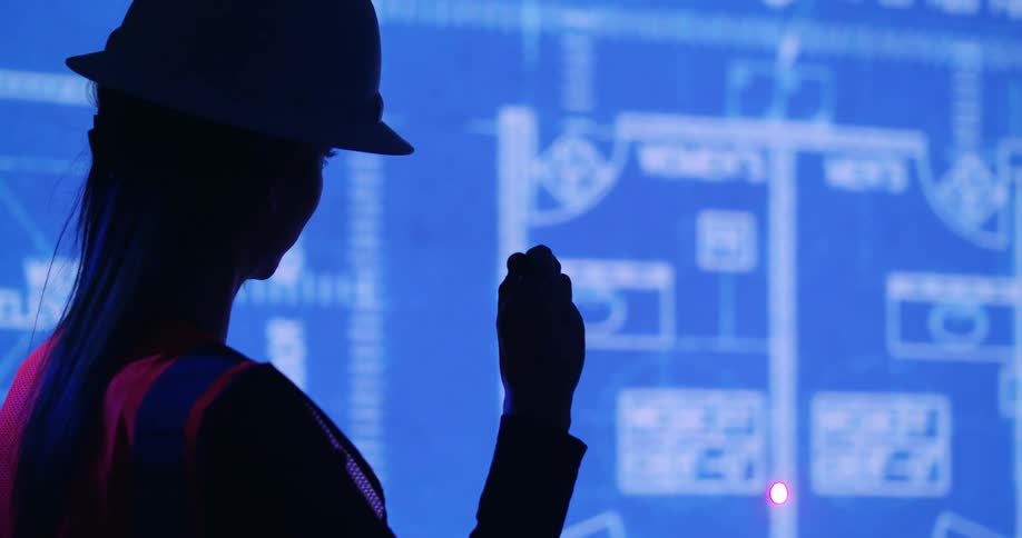 A woman wearing a hard hat and an orange safety vest uses a laser pointer on a large illuminated blueprint displayed on a screen.