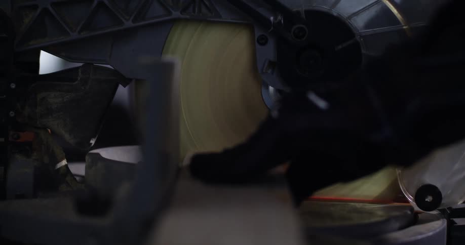 View from a piece of wood of a hand holding the wood while pulling down the handle of the rotating blade of a miter saw to cut a piece of wood.