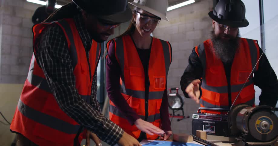 A female and two male construction engineers analyze project blueprints together on a table at a building site while a welder sends sparks flying in the background.